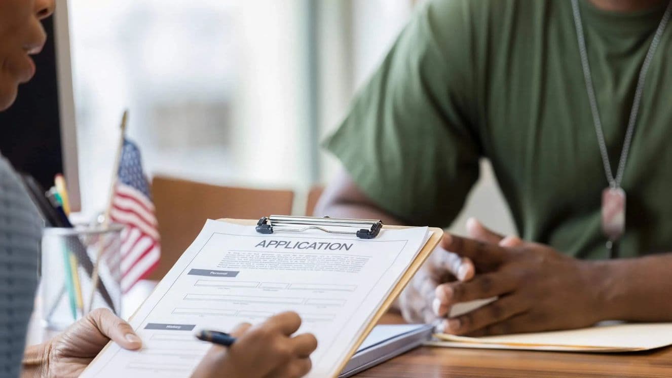 Person reviewing an application form on a clipboard while speaking with a military service member seated across a desk, with a small American flag visible in the background.